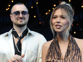 Sandra Kubicka i Aleksander Baron w modnych ubraniach na tle delikatnego oświetlenia, uśmiechają się do aparatu.