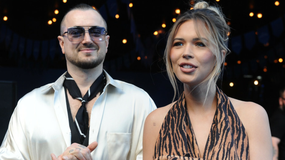 Sandra Kubicka i Aleksander Baron w modnych ubraniach na tle delikatnego oświetlenia, uśmiechają się do aparatu.