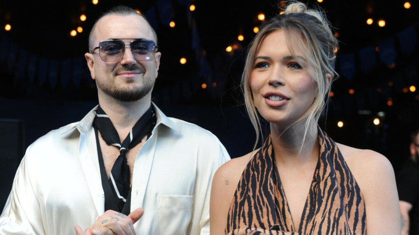 Sandra Kubicka i Aleksander Baron w modnych ubraniach na tle delikatnego oświetlenia, uśmiechają się do aparatu.
