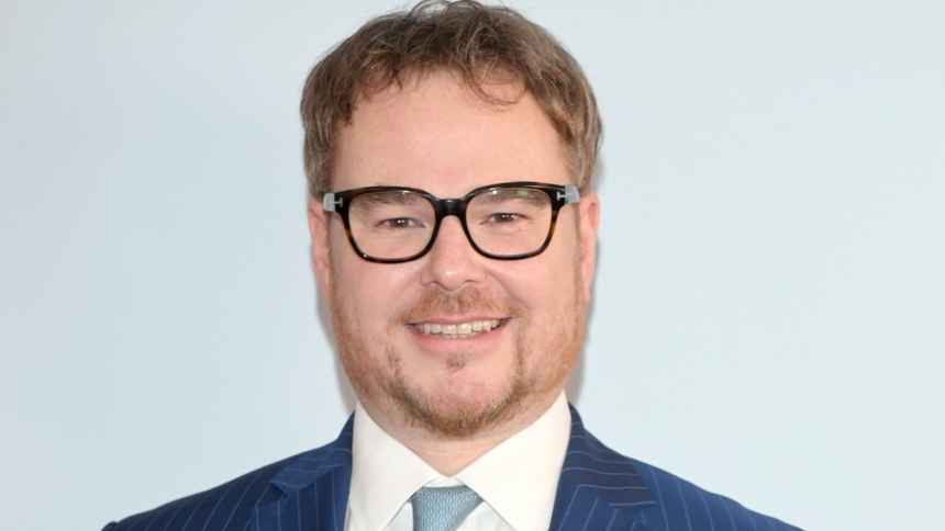 Uśmiechnięty Jan Lubomirski-Lanckoroński w okularach, w niebieskim garniturze i jasnej koszuli, na jasnym tle.