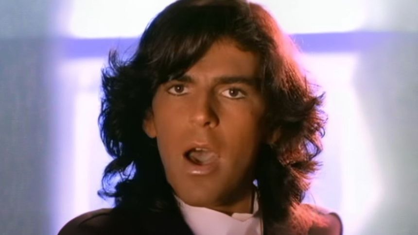 Mężczyzna z długimi, ciemnymi włosami śpiewa. Tło jasne.