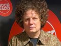 Mężczyzna z kręconymi włosami, w brązowej koszuli, stoi na tle logo Rock Radio.