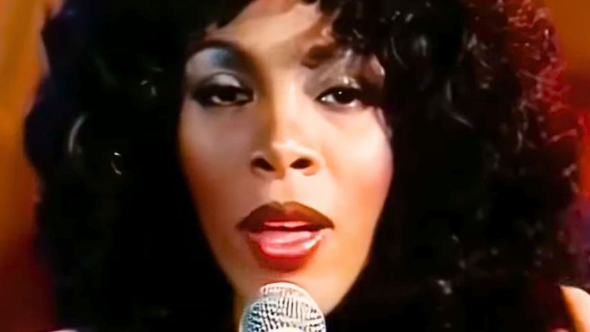 Na zdjęciu artystka z mikrofonem, z długimi kręconymi włosami i wyrazistym makijażem.