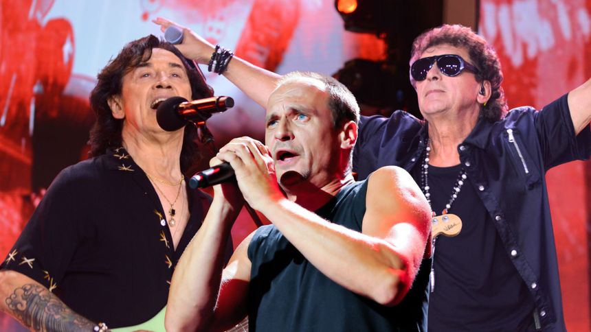 Trzech mężczyzn na scenie, śpiewających. W tle kolorowe światła, rockowy klimat koncertu.