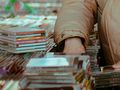 Osoba w beżowej kurtce przeszukuje stos płyt CD na targu. W tle widać inne płyty.