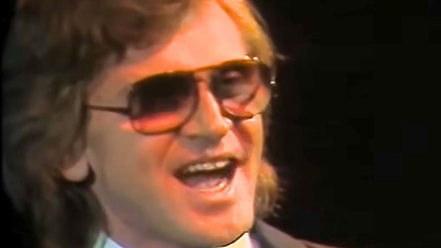 Mężczyzna w dużych okularach przeciwsłonecznych, śpiewający z uśmiechem na scenie.