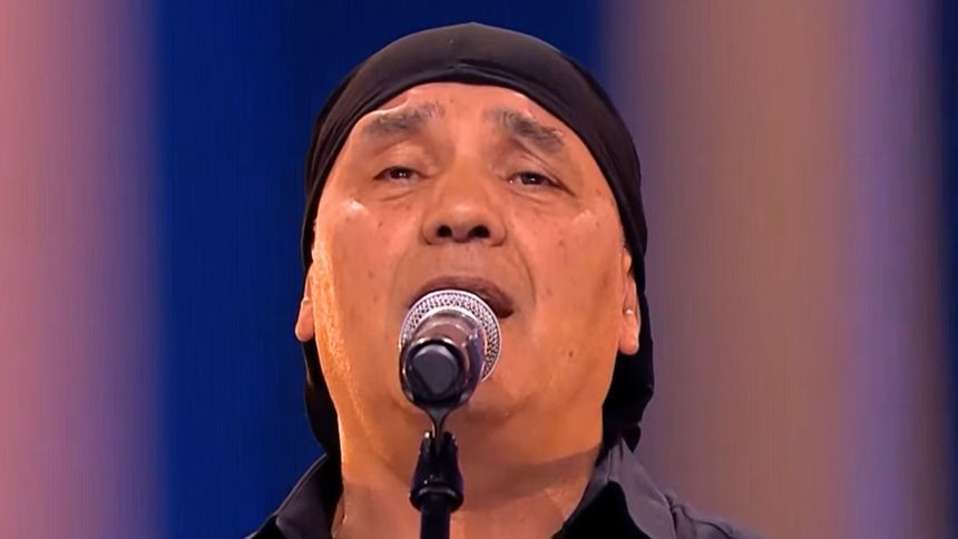 Mężczyzna w czarnym t-shircie i chustce, śpiewający do mikrofonu na scenie.