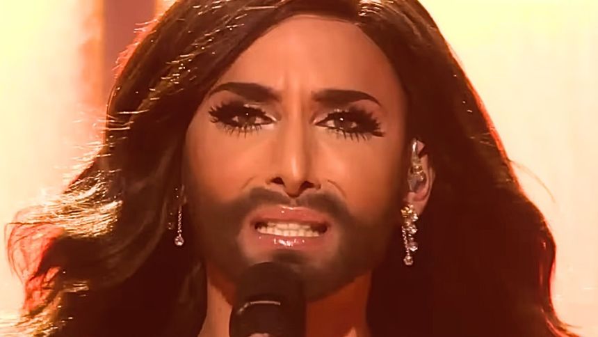 Na zdjęciu artysta śpiewający, z długimi włosami, makijażem i brodą. Intensywne emocje.