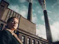 Kobieta w płaszczu stoi przed fabryką z dymiącymi kominami w tle, patrząc w dal.
