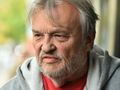 Mężczyzna z siwymi włosami i brodą, w szarej bluzie, z poważnym wyrazem twarzy