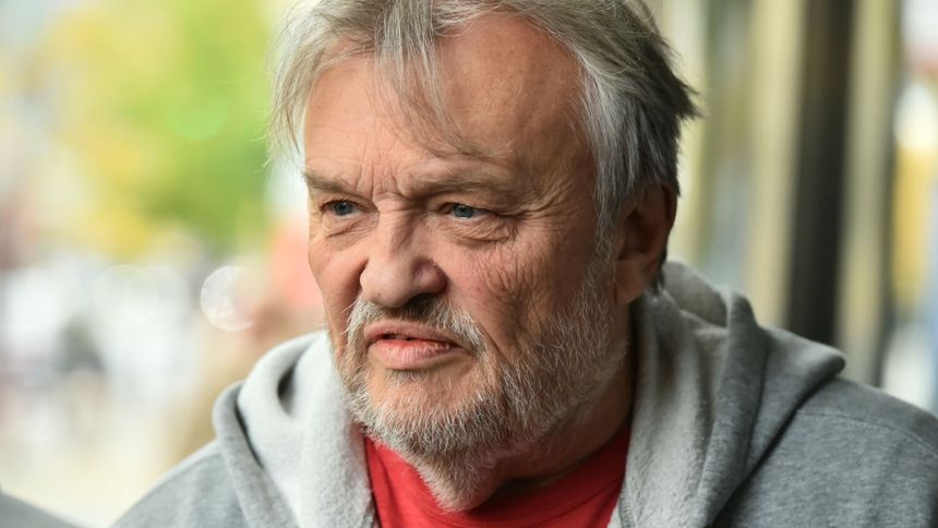 Mężczyzna z siwymi włosami i brodą, w szarej bluzie, z poważnym wyrazem twarzy