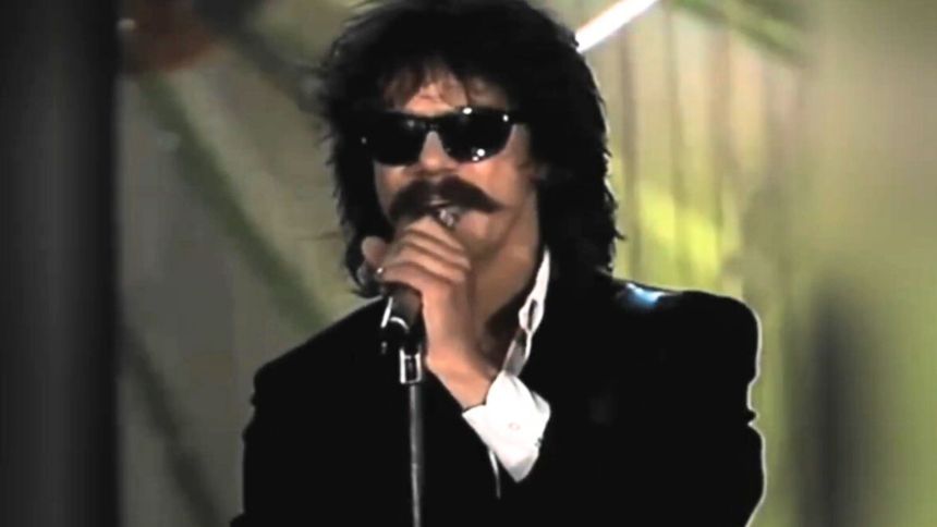 Mężczyzna w ciemnych okularach, ubrany na czarno, śpiewa do mikrofonu, intensywne oświetlenie w tle.