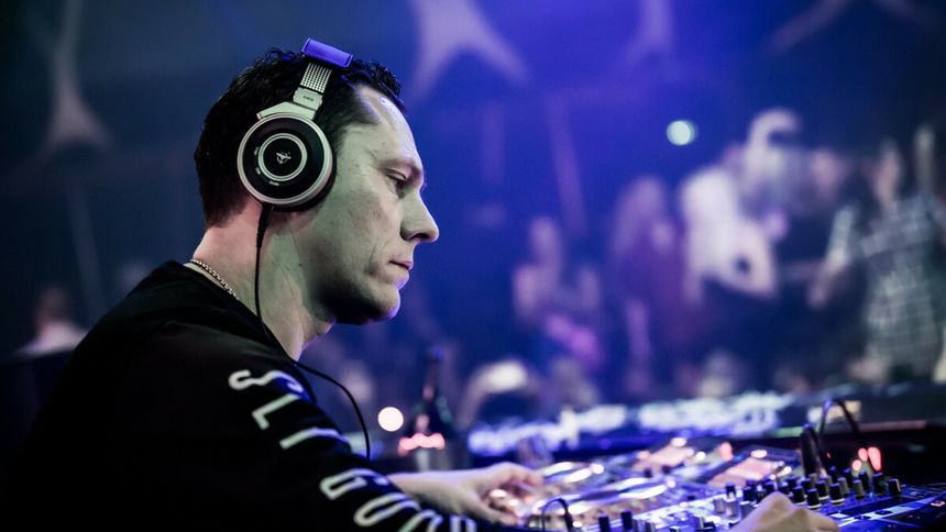 DJ Tiesto w słuchawkach przy konsoli, grający muzykę na imprezie w nocnym klubie. Tłum w tle.
