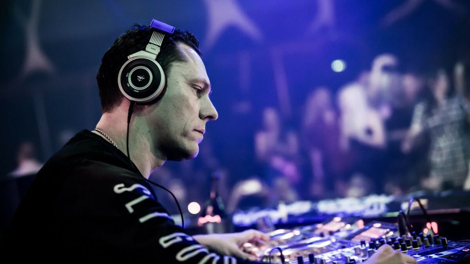 DJ Tiesto w słuchawkach przy konsoli, grający muzykę na imprezie w nocnym klubie. Tłum w tle.