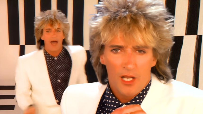 Dwaj mężczyźni w jasnych garniturach, z blond włosami, na tle w czarno-białe paski.