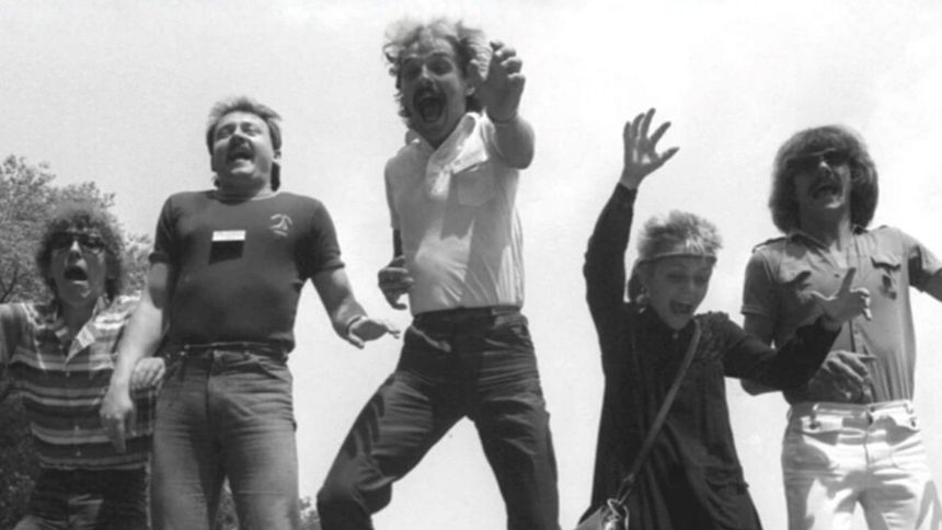 Grupa pięciu osób skacze i wyraża radość w plenerze, w tle drzewa i niebo