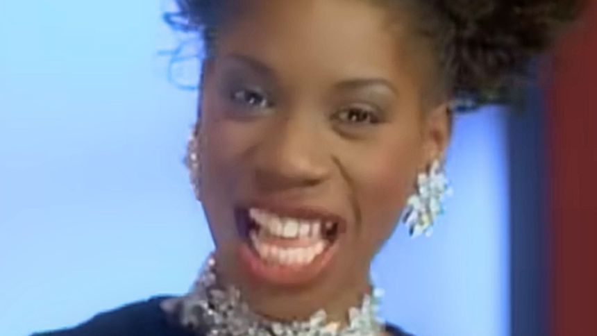 Osoba uśmiechnięta, w błyszczącej biżuterii, z kręconymi włosami, na jasnym tle.