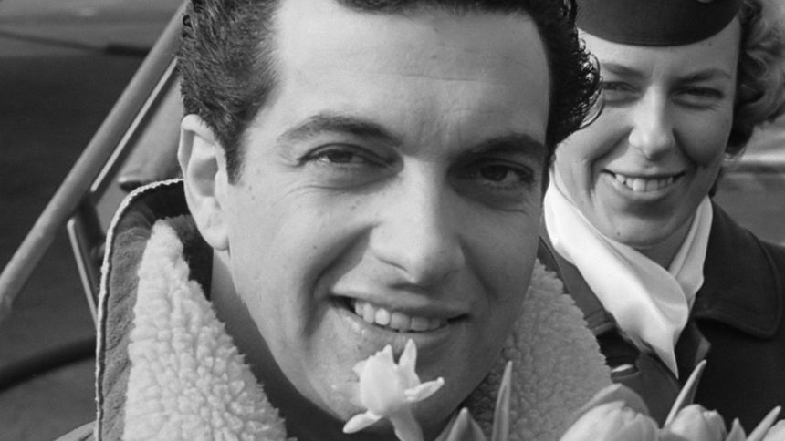 Mężczyzna z bukietem tulipanów, uśmiechnięty, w kurtce z kożuszkiem; za nim kobieta w mundurze.