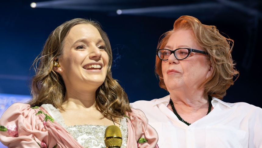 Na zdjęciu dwie kobiety, jedna z mikrofonem, uśmiechnięta, druga z okularami, poważna