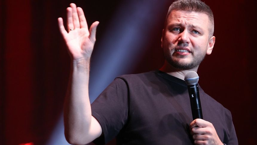 Mateusz Socha w czarnej koszulce trzyma mikrofon i gestykuluje, stojąc na scenie.