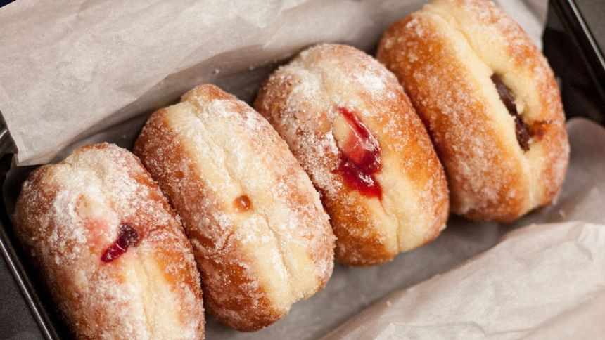 Cztery pączki na papierze, jeden z dżemem, reszta z nadzieniem kremowym, posypane cukrem pudrem.