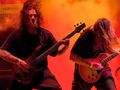 Muzycy grają na scenie, jeden z nich na basie, drugi na gitarze. Intensywne oświetlenie.