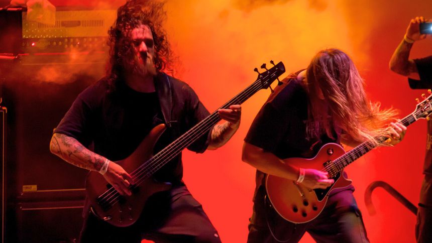 Muzycy grają na scenie, jeden z nich na basie, drugi na gitarze. Intensywne oświetlenie.