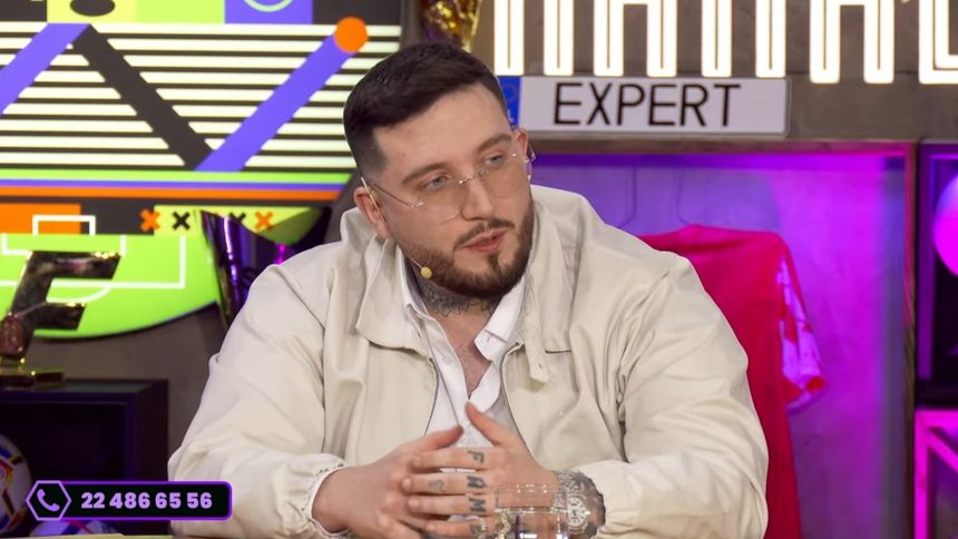 Michał „Boxdel” Baron w jasnej kurtce i okularach, siedzi przy stole, gestykulując podczas rozmowy.
