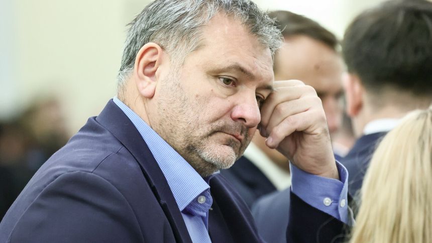 Mężczyzna w garniturze, zamyślony, dotyka czoła, w tle inni ludzie.