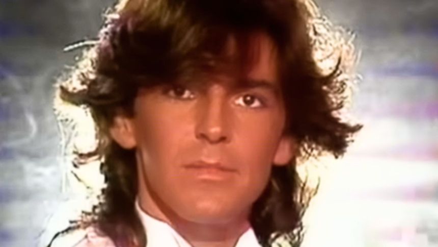 Mężczyzna z długimi włosami, w białej marynarce, patrzy w obiektyw na tle rozmytego tła.