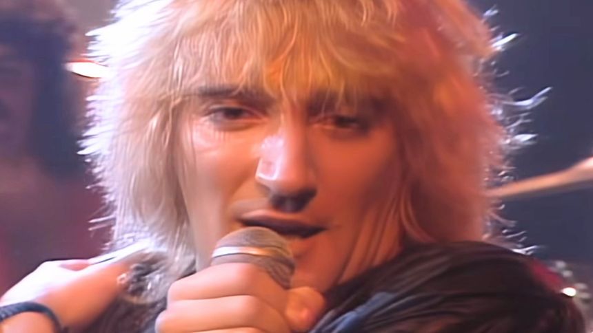 Mężczyzna z długimi, blond włosami, śpiewający do mikrofonu na scenie w blasku świateł.