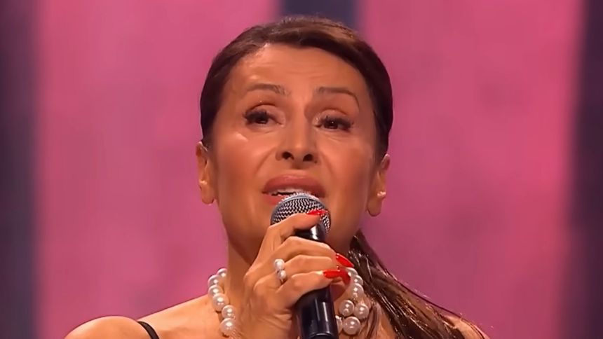 Kobieta z mikrofonem na scenie, w eleganckiej biżuterii, w tle różowa tapeta.