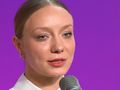 Kobieta z jasnymi włosami, ubrana w białą koszulę, mówi do mikrofonu na tle fioletowym.