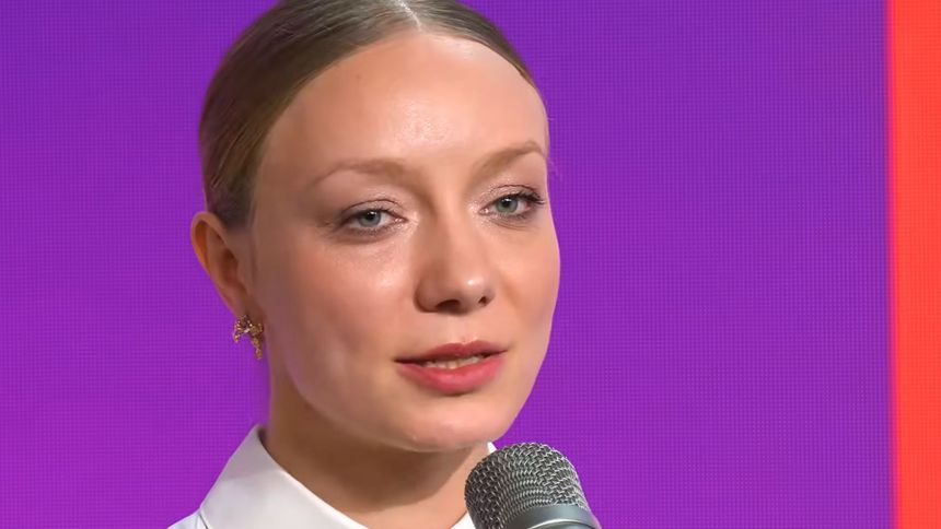 Kobieta z jasnymi włosami, ubrana w białą koszulę, mówi do mikrofonu na tle fioletowym.