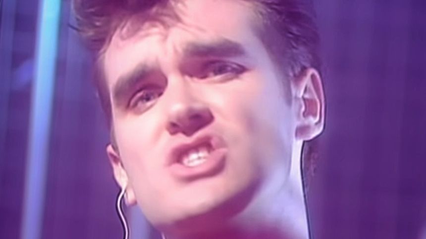 Mężczyzna z krótkimi włosami, śpiewający na tle fioletowego światła. Wyraża emocje.