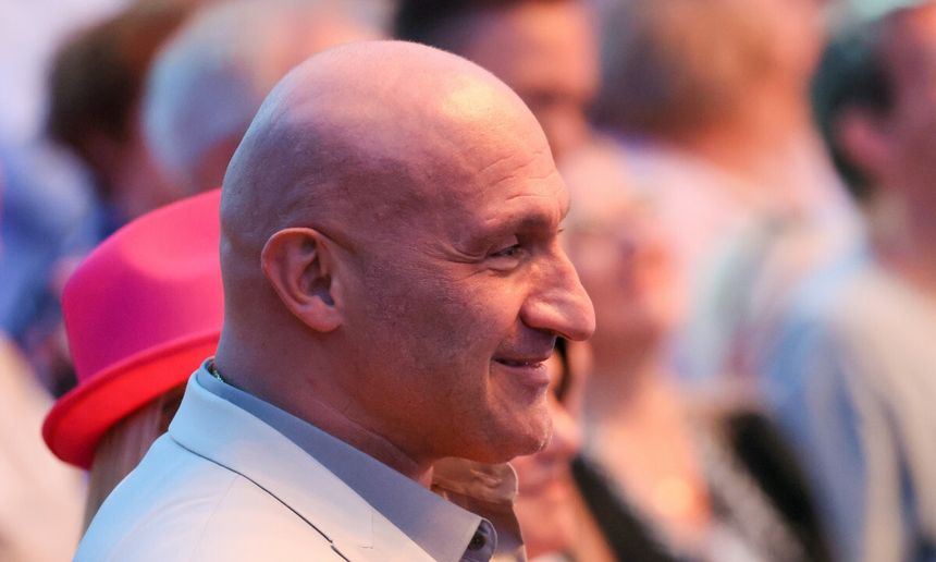 Na zdjęciu Marcin Najman z łysą głową, uśmiechający się, w otoczeniu ludzi i rozmytym tłem.
