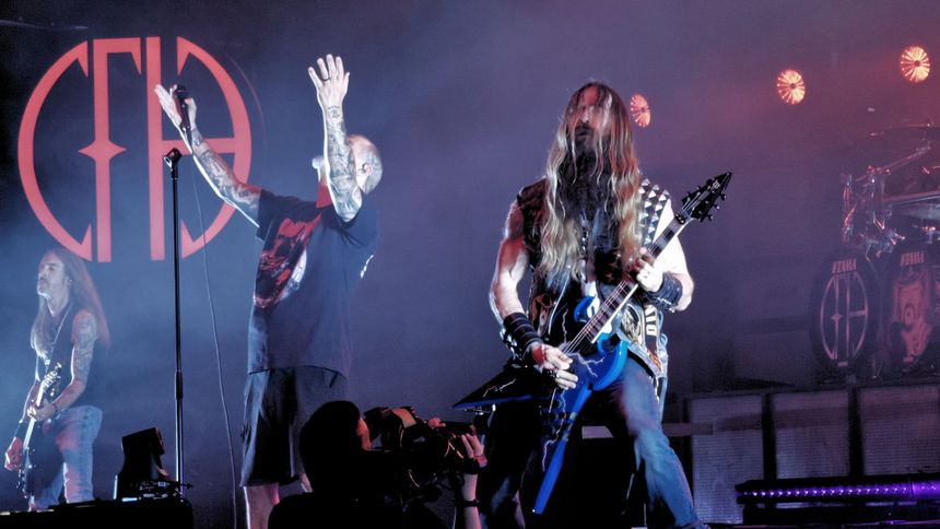 Trzech muzyków na scenie, jeden z mikrofonem, dwóch gra na gitarach, intensywne oświetlenie