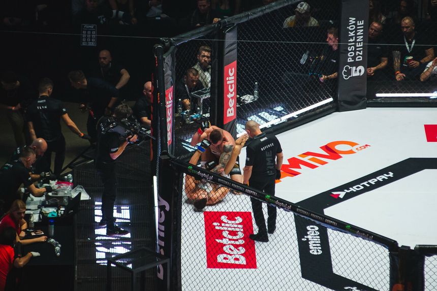 FAME MMA: zawodnik w akcji na ringu, sędzia ocenia sytuację, grupa ludzi w tle.