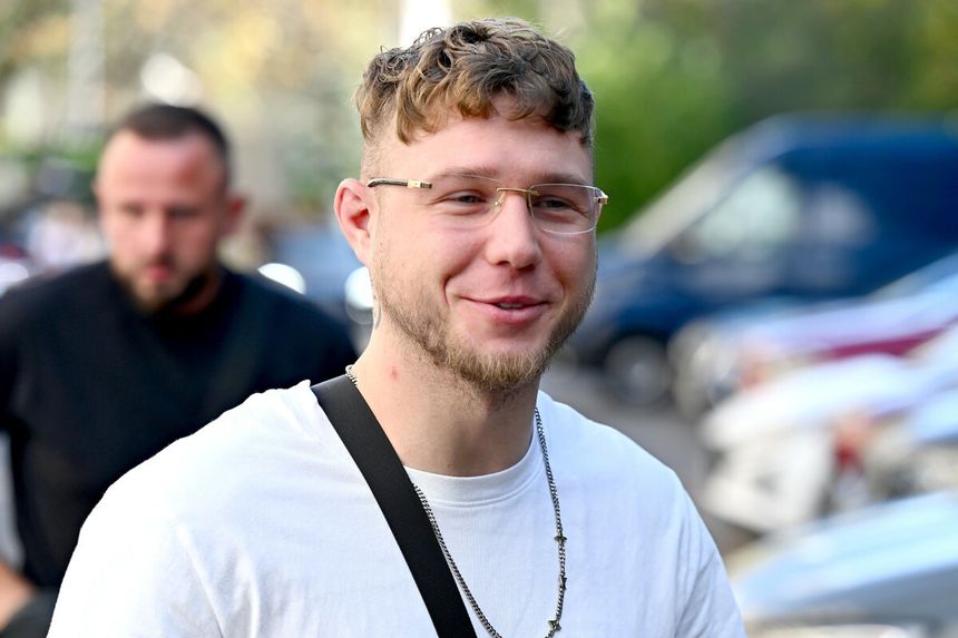 Jakub „Kubańczyk” Flas z okularami, w białej koszulce, uśmiecha się na tle miasta.