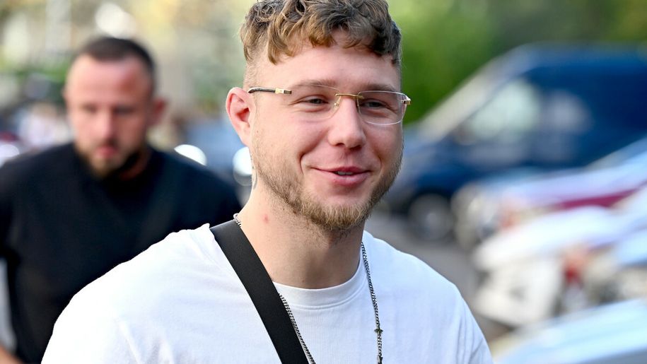 Jakub „Kubańczyk” Flas z okularami, w białej koszulce, uśmiecha się na tle miasta.