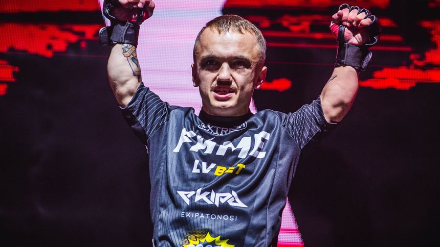 Mini Majk w sportowym stroju FAME, unosi ręce w geście zwycięstwa na tle intensywnego światła.