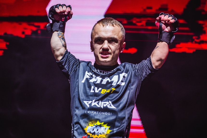 Mini Majk w sportowym stroju FAME, unosi ręce w geście zwycięstwa na tle intensywnego światła.