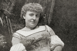 Frances Hodgson Burnett. Kobieta w długiej sukni, z lokami, siedząca na krześle, otoczona roślinami. Czarno-białe zdjęcie.