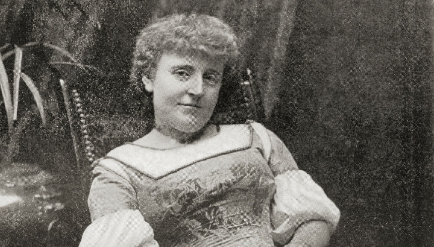 Frances Hodgson Burnett. Kobieta w długiej sukni, z lokami, siedząca na krześle, otoczona roślinami. Czarno-białe zdjęcie.