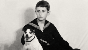 Chłopiec w marynarskim ubraniu siedzi z psem na tle jasnej ściany.