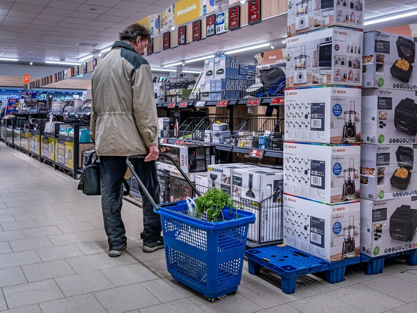 Mężczyzna w kurtce z wózkiem na zakupy, stoi w sklepie przy półkach z produktami AGD.