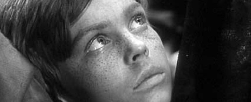 Młody chłopiec z jasnymi włosami i piegami, patrzy w górę z zamyśloną miną.