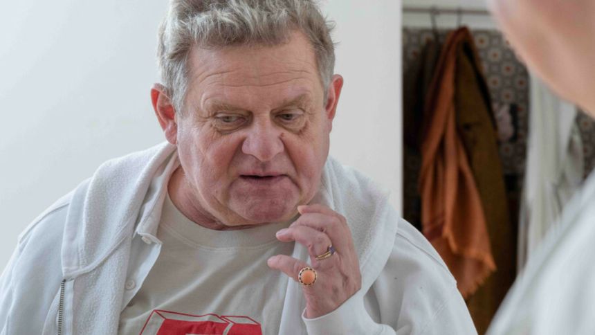 Mężczyzna w białej bluzie z kapturem, z krótkimi, siwymi włosami, dotyka brody, głęboko myśląc