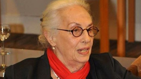 Kobieta z siwymi włosami i okularami, w czarnym stroju z czerwoną chustką na szyi.