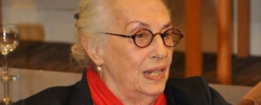Kobieta z siwymi włosami i okularami, w czarnym stroju z czerwoną chustką na szyi.